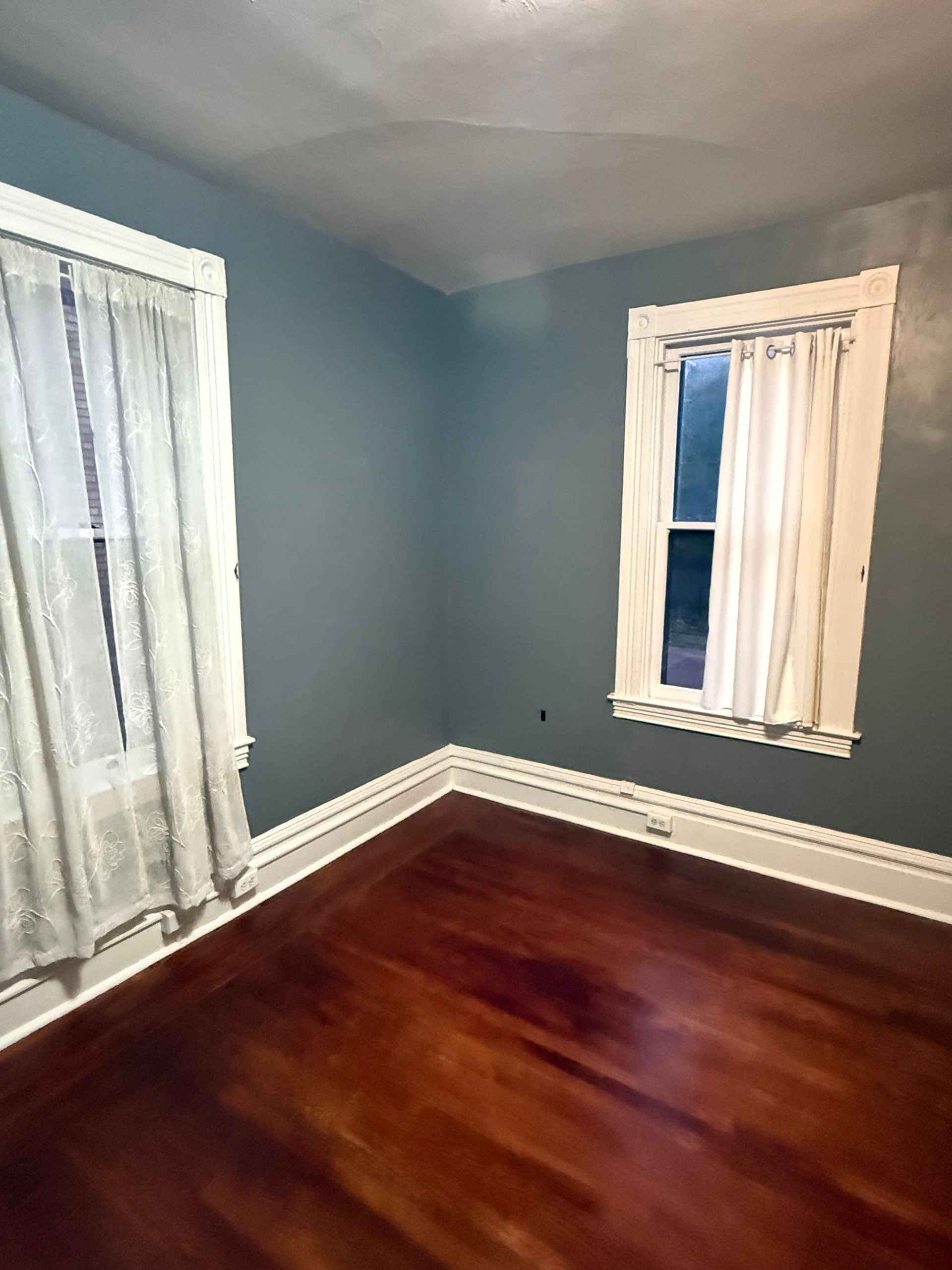 Empty blue room with hardwood floor, white trim, and two curtained windows.