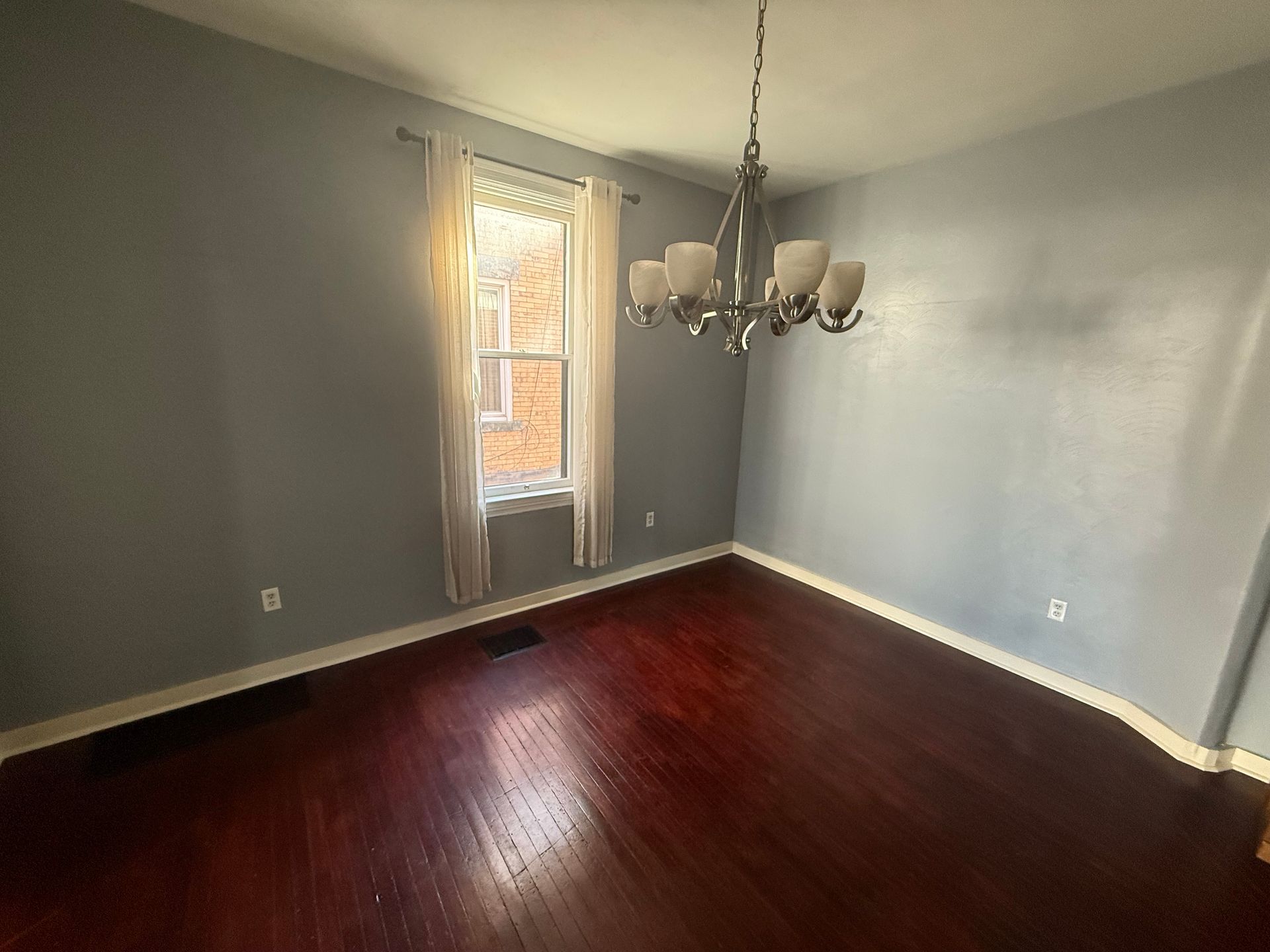 Empty room with dark hardwood floors, gray walls, a window, and a chandelier hanging from the ceiling