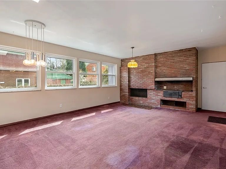 Empty living room with pink carpet, brick fireplace, and large windows letting in daylight