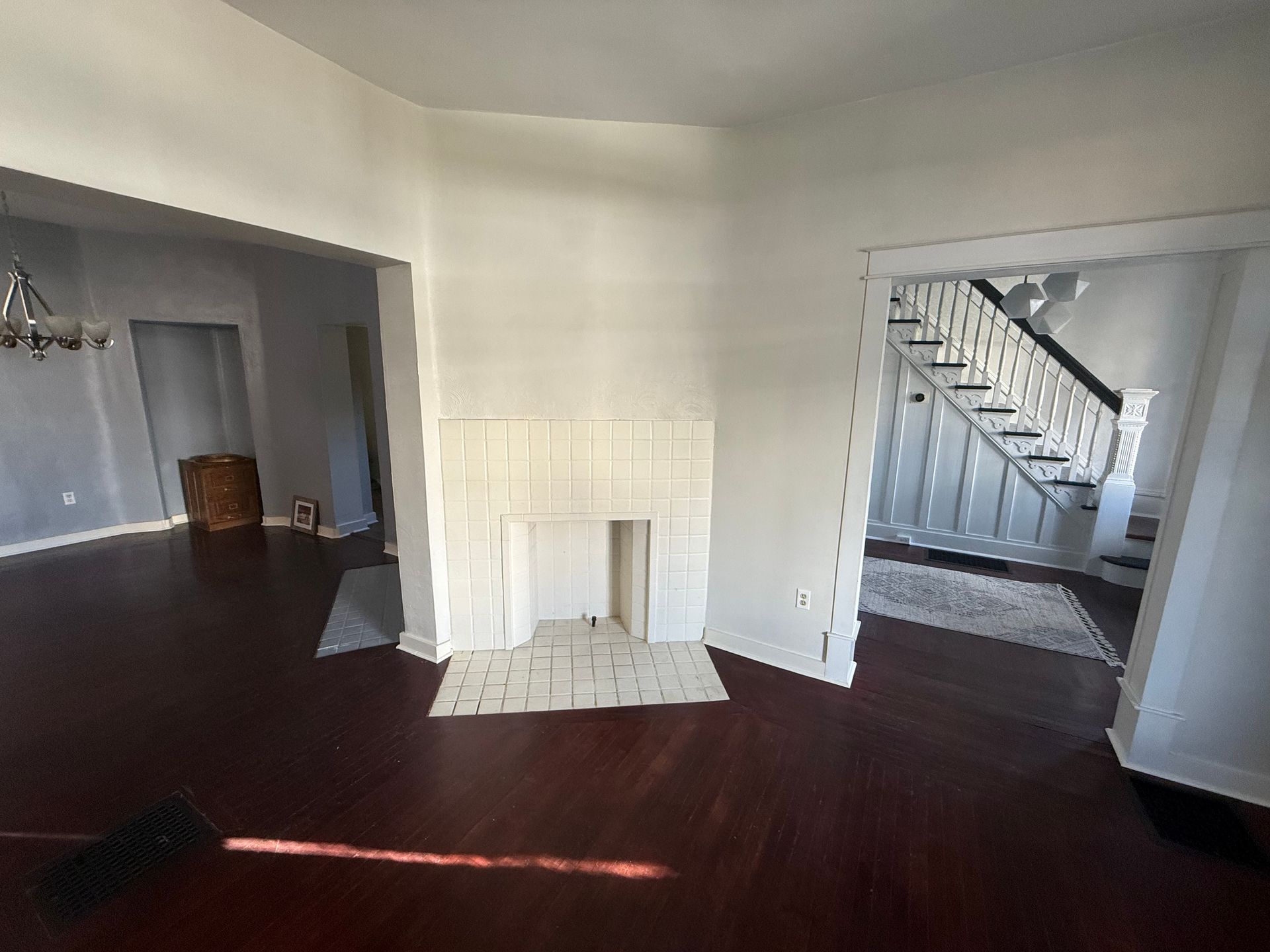 Empty living room with dark wood floors, a white fireplace, and a staircase visible through a doorway.
