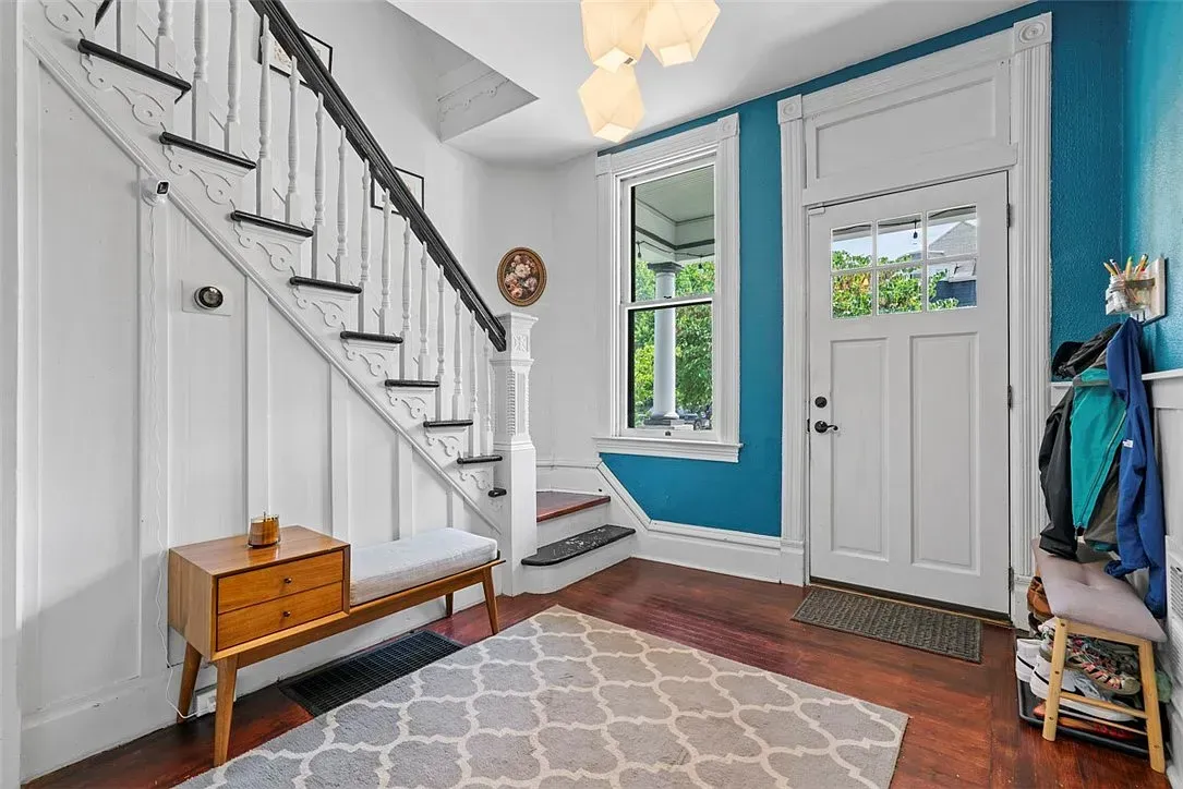 Bright entryway with white staircase, blue accent wall, double doors, bench, and patterned rug