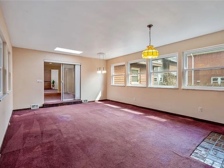 Empty beige living room with burgundy carpet, windows, mirrored closet doors, and a yellow pendant light