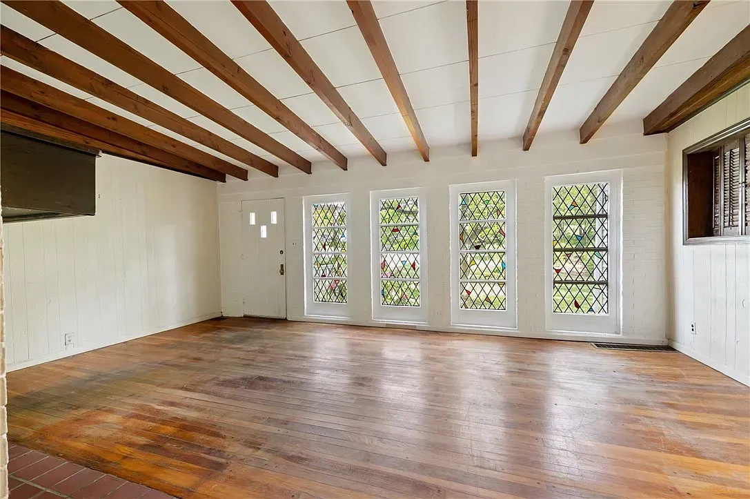 Empty room with wooden beams, hardwood floors, and tall windows overlooking greenery