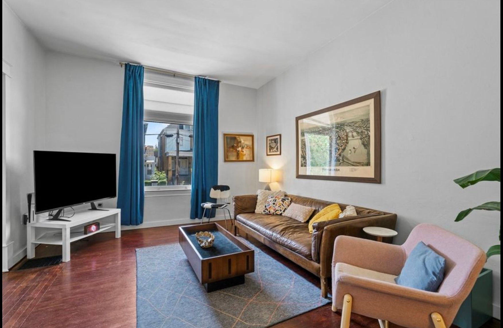 Bright living room with blue curtains, brown sofa, TV, coffee table, and framed wall art