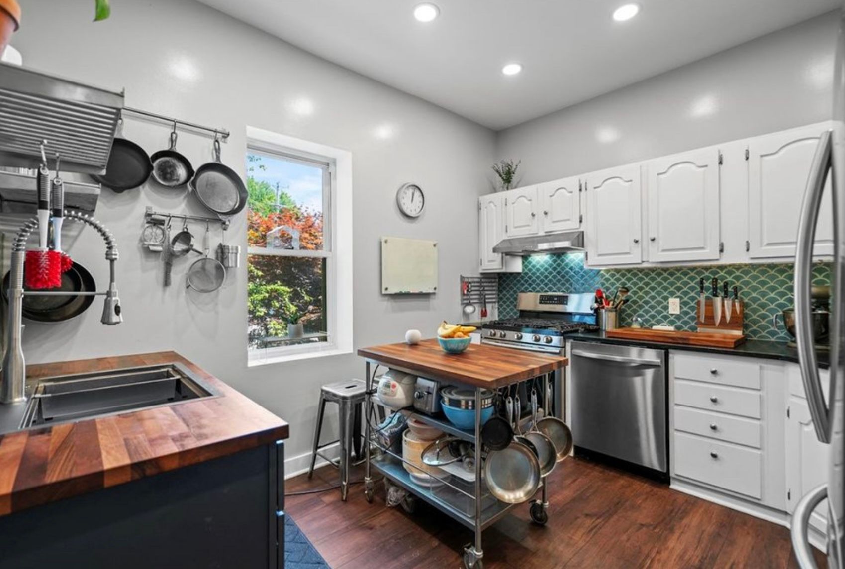 Modern kitchen with white cabinets, stainless appliances, wood island, and hanging pots by a bright window