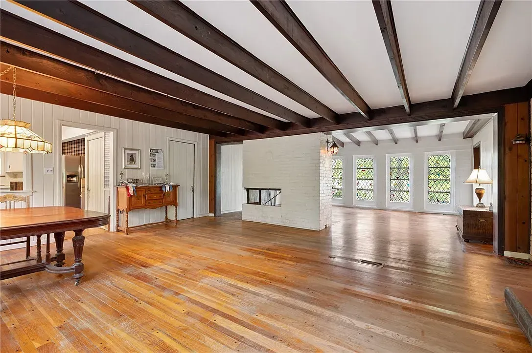 Spacious empty living room with hardwood floors, exposed beams, and large windows overlooking greenery