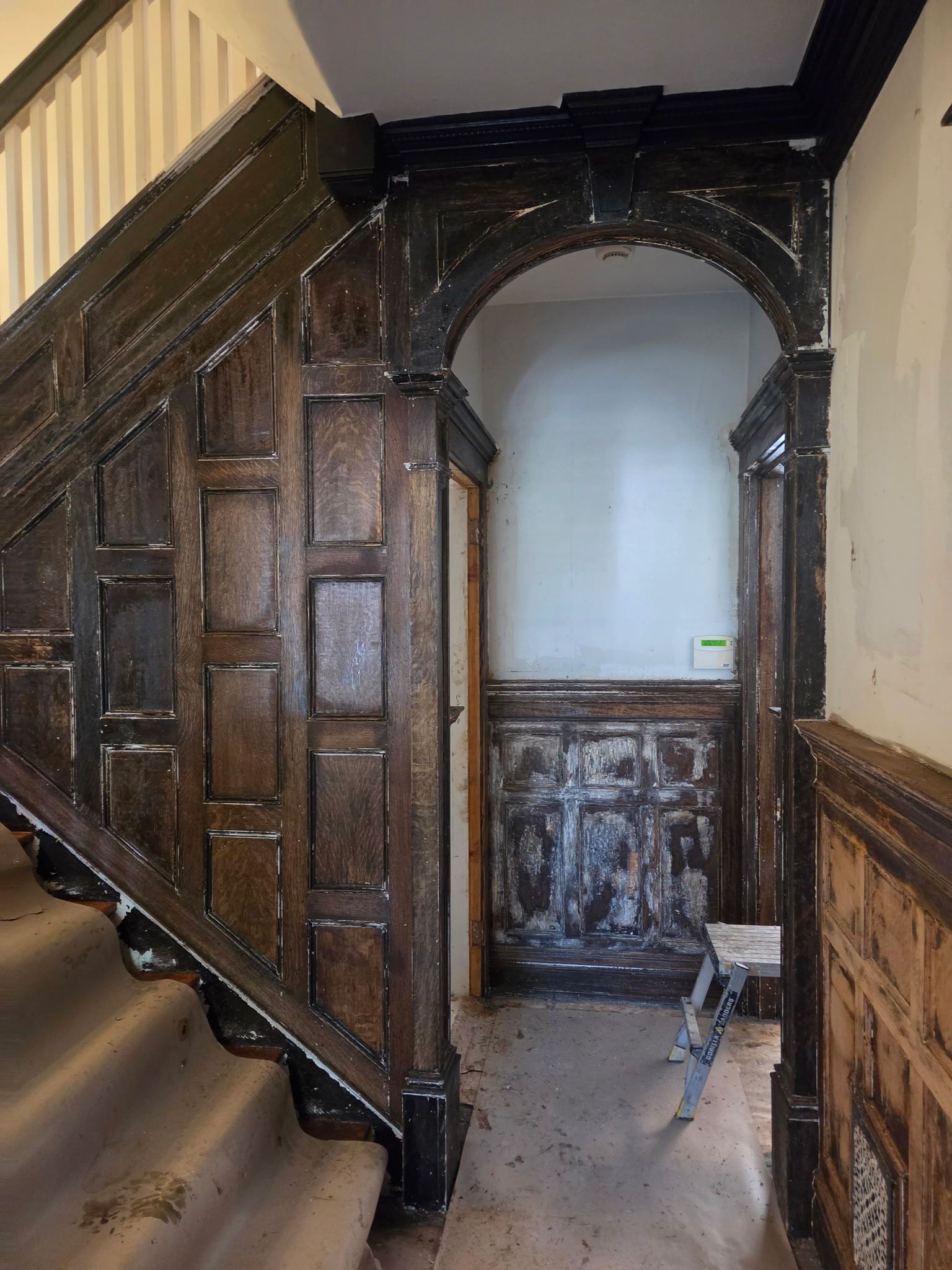 Dark wooden staircase with ornate paneling and arched doorway in a narrow hallway.