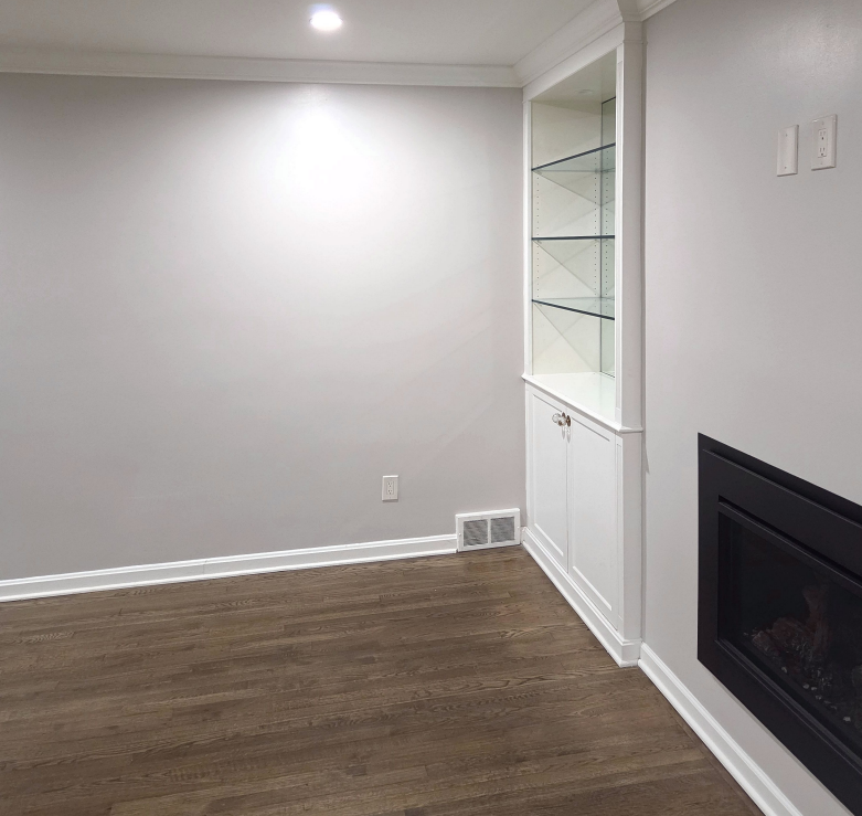 Empty living room with gray walls, wood floor, built-in shelves, and a black fireplace.
