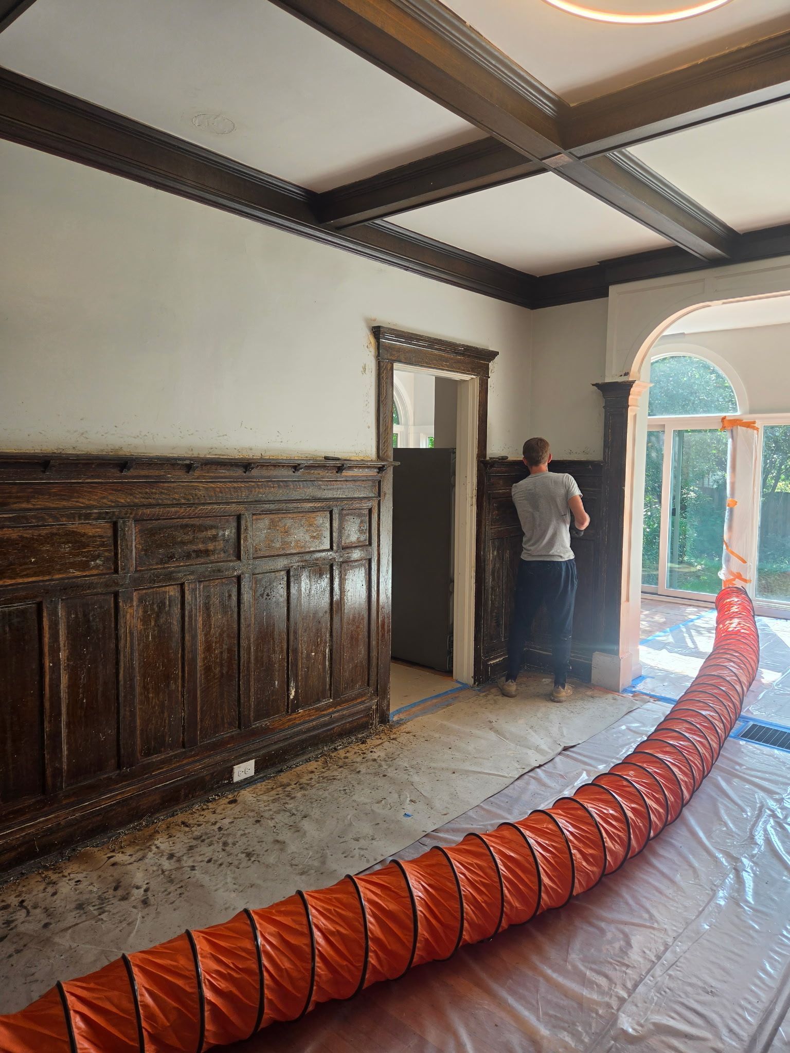 Worker standing in a room with dark wood trim, orange ventilation duct on the floor, and daylight through a glass door.