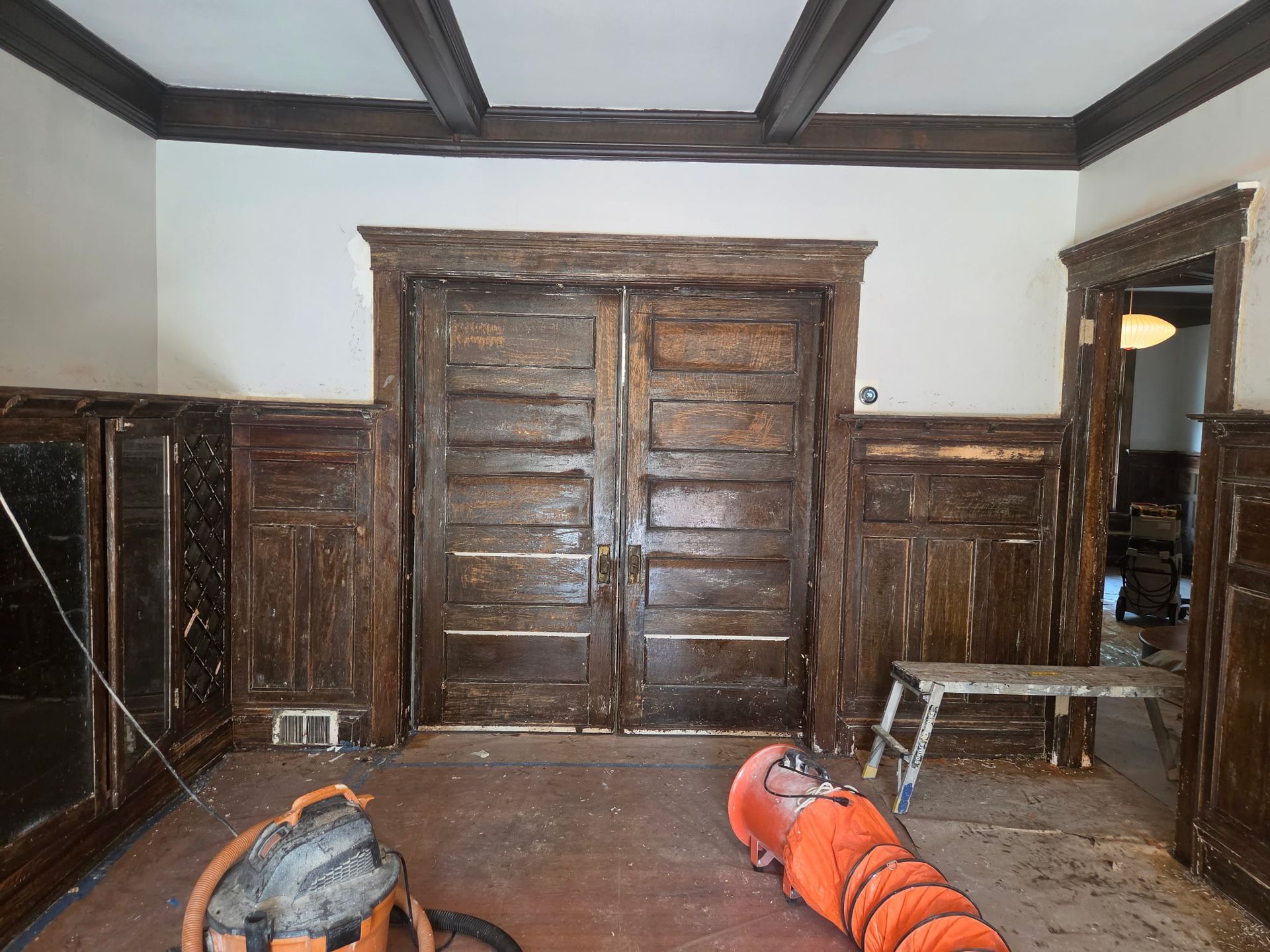 Wood-paneled room with double wooden doors, benches, and two orange vacuum cleaners on the floor.