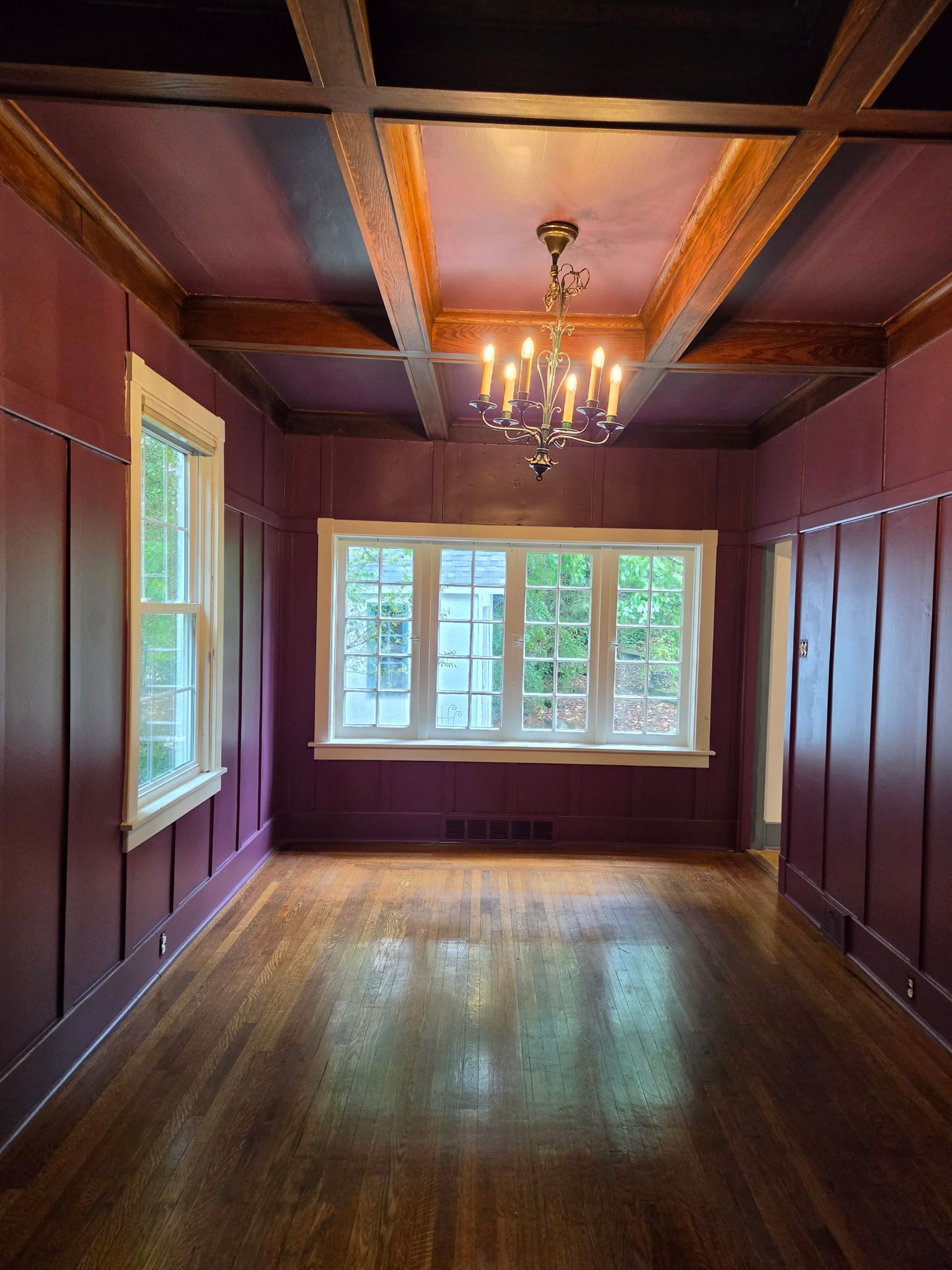 Empty room with dark wood floors, purple walls, exposed beams, chandelier, and a large window at the far end.