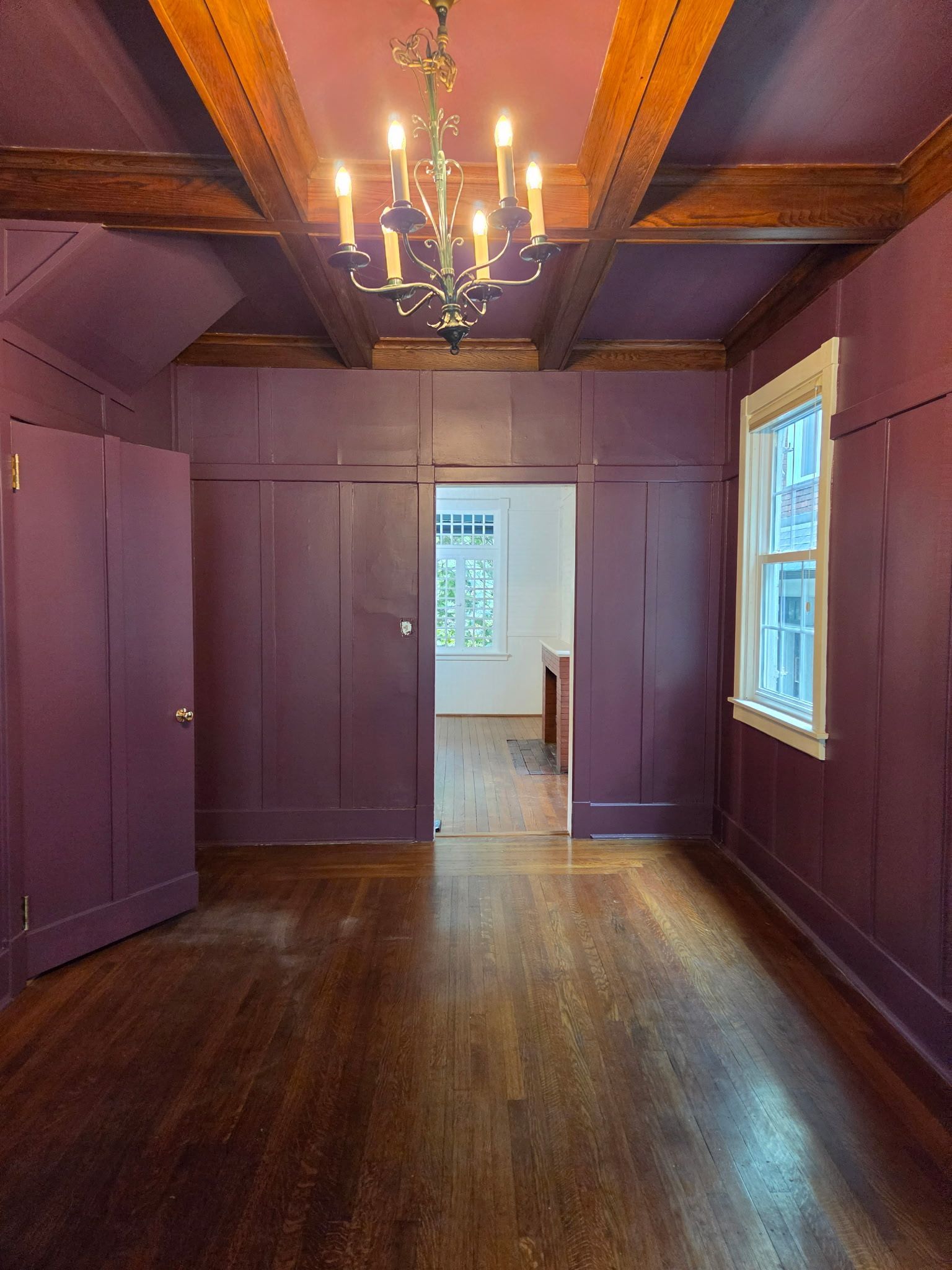 Empty purple room with wood floors, chandelier, and doorway to a bright adjoining room.