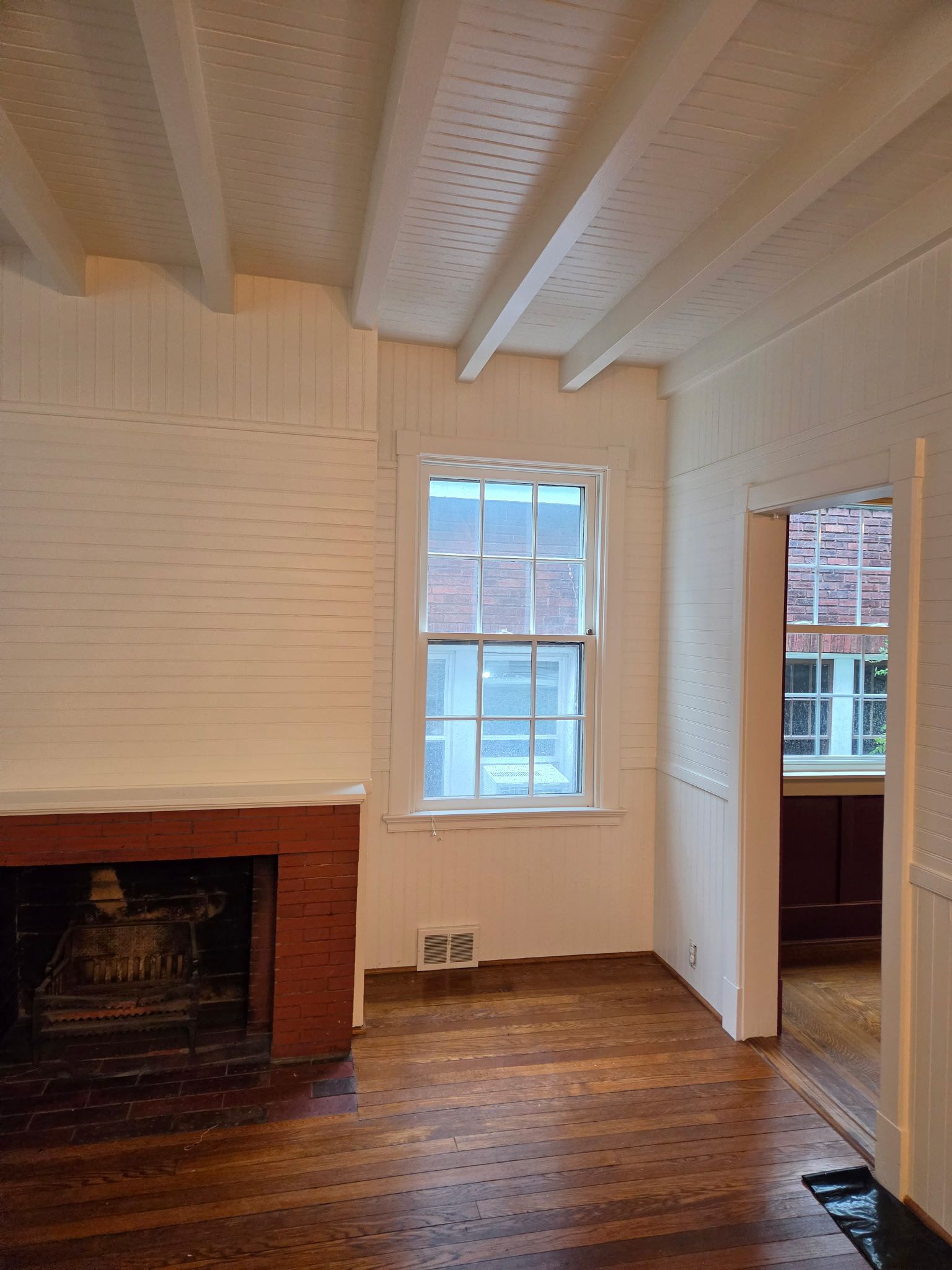 White-painted room with a brick fireplace, hardwood floor, and a window beside an open doorway.