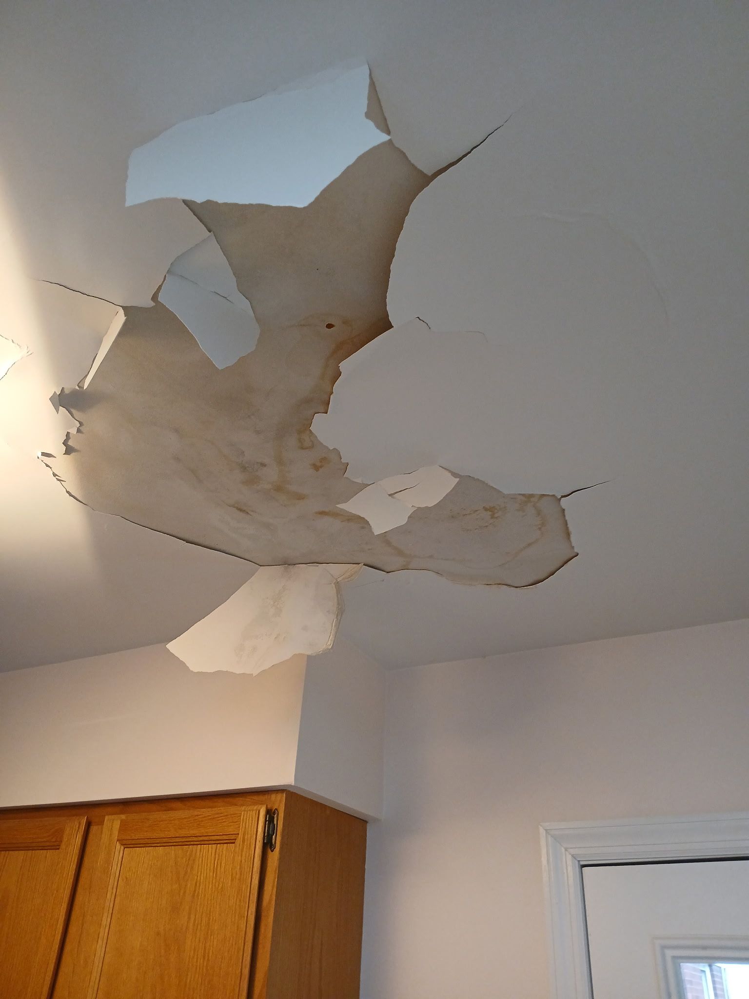 Damaged ceiling with a large hole and water stains above kitchen cabinets.