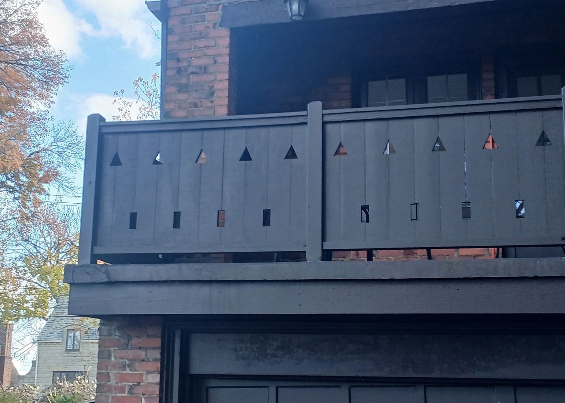Black balcony railing with cut-out shapes on a brick building, view of the sky and trees.