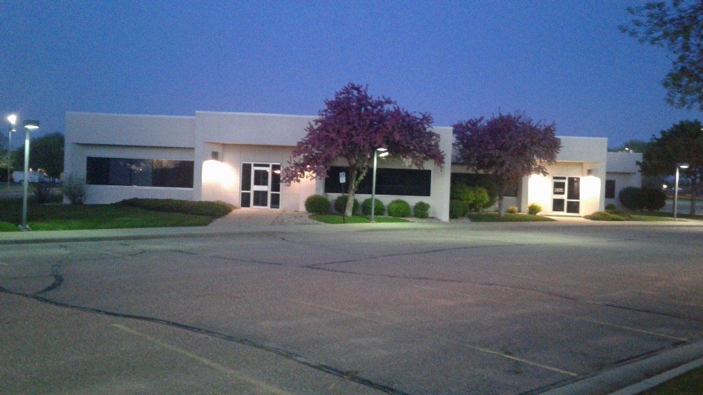 Modern, white building with dark windows and doors, lit by streetlights. Purple-leaved trees in front.