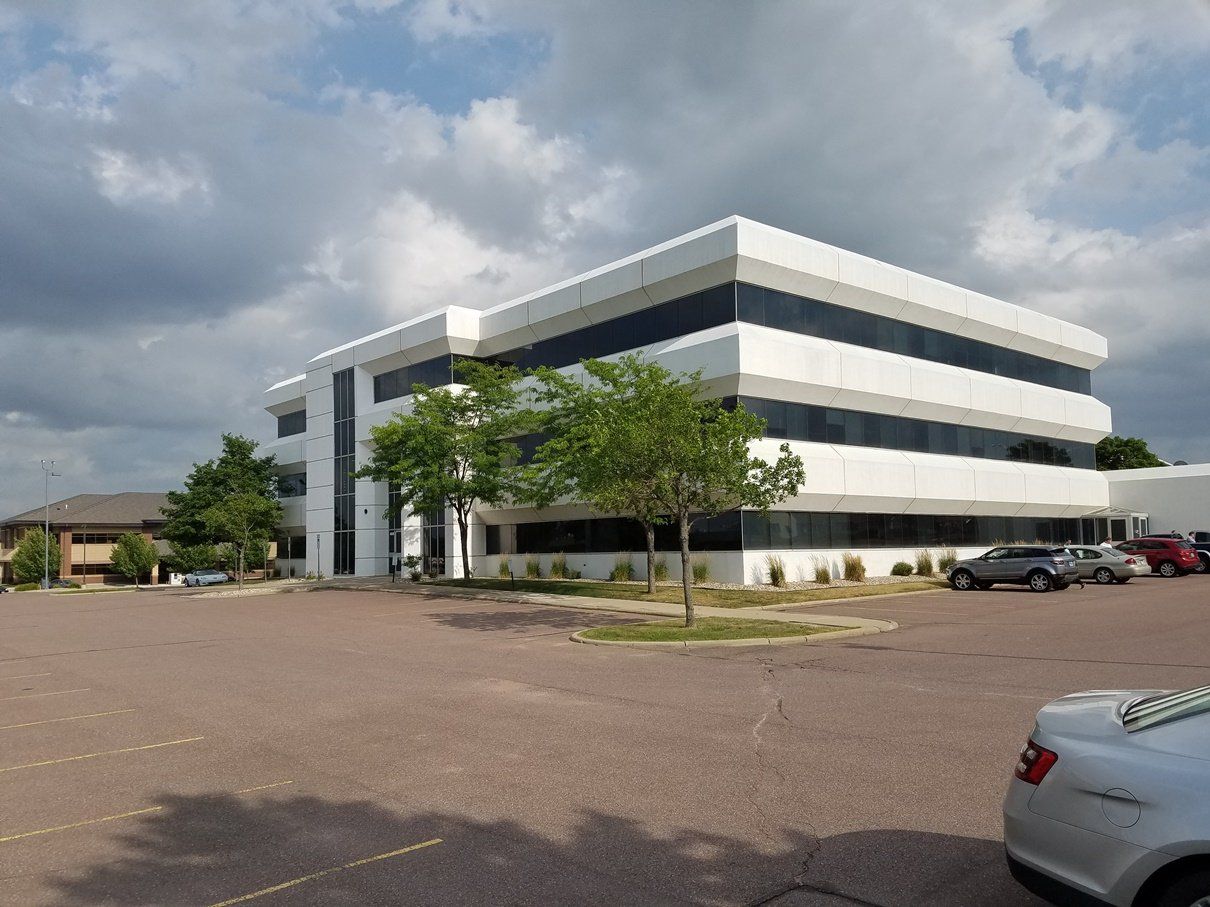 White office building with black-framed windows, surrounded by a paved parking lot with cars and some trees.