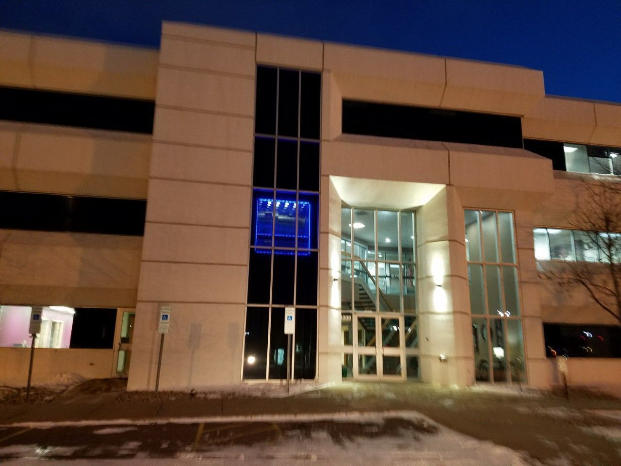 Modern building entrance with glass doors and windows, illuminated at night. Snow on the ground.