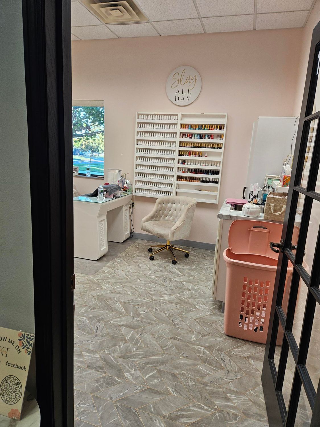 Interior of a nail salon, pink walls, white shelving with polish, chair, laundry basket, open doorway.