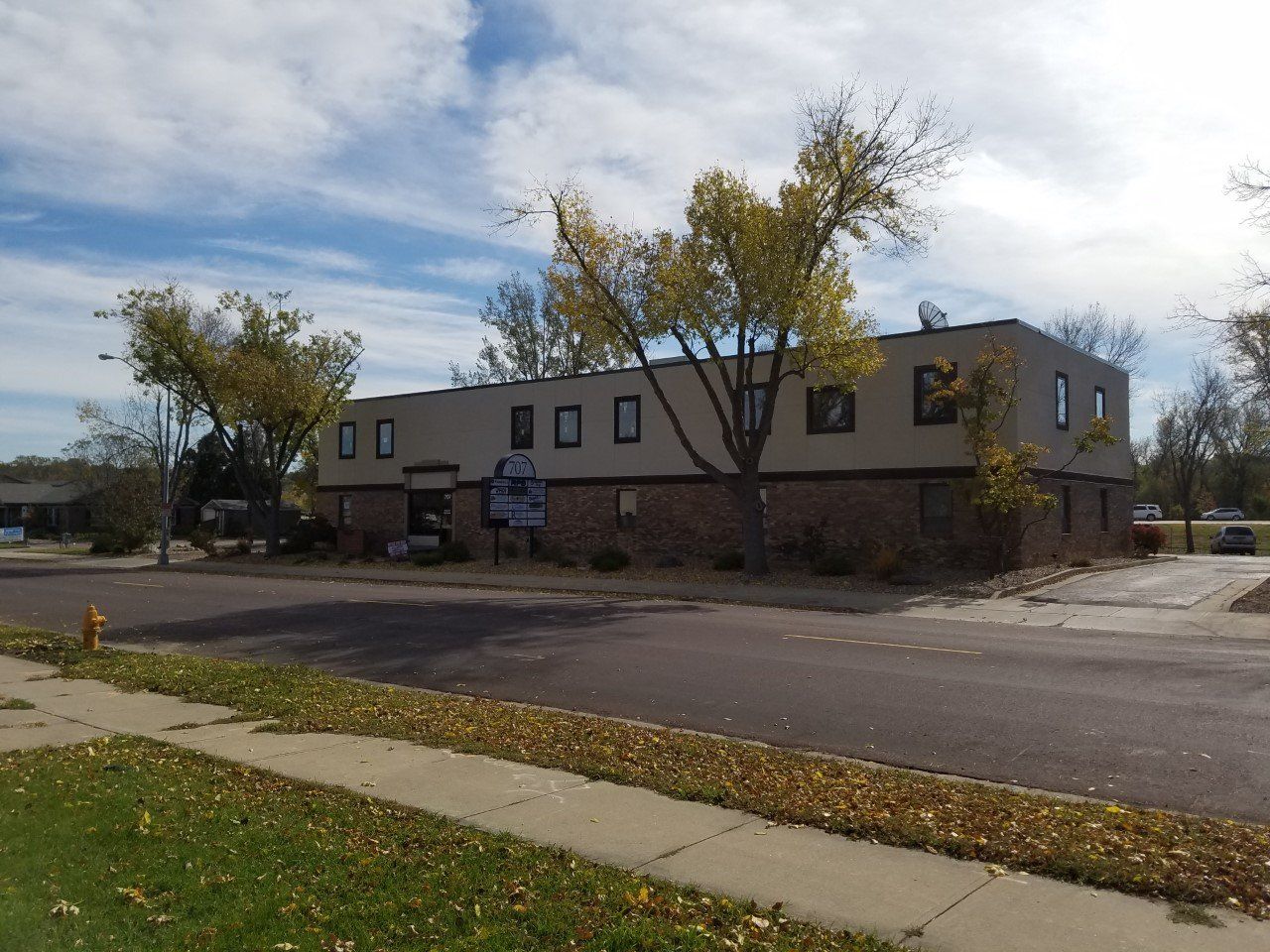 Two-story beige brick building with dark windows and trees, viewed from a street.