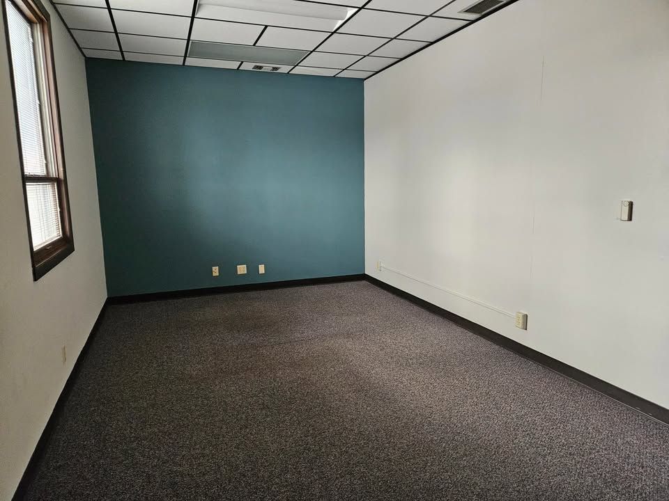 Empty office room with gray carpet, teal accent wall, white walls, and a window.