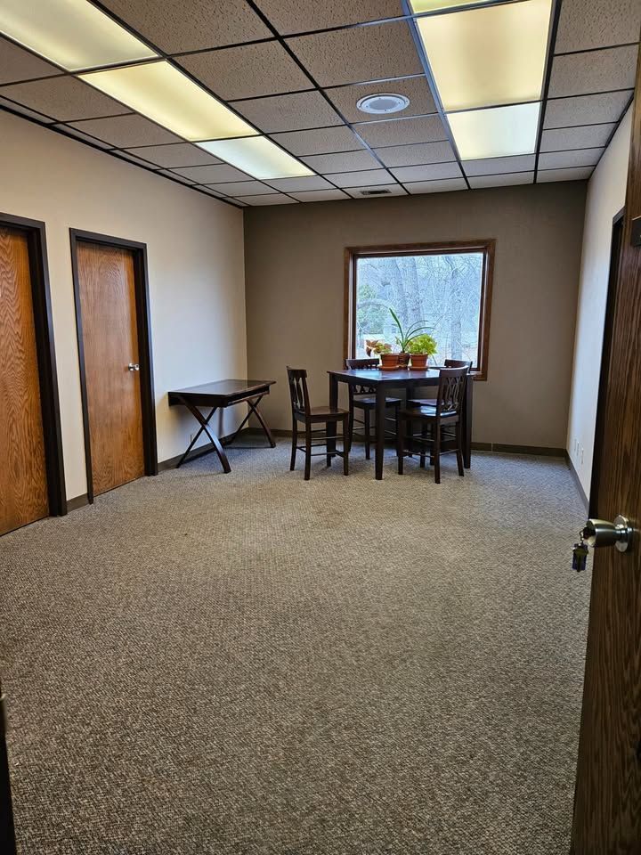 Empty office room with table, chairs, window, and two doors. Brown carpet and walls. Fluorescent lighting.