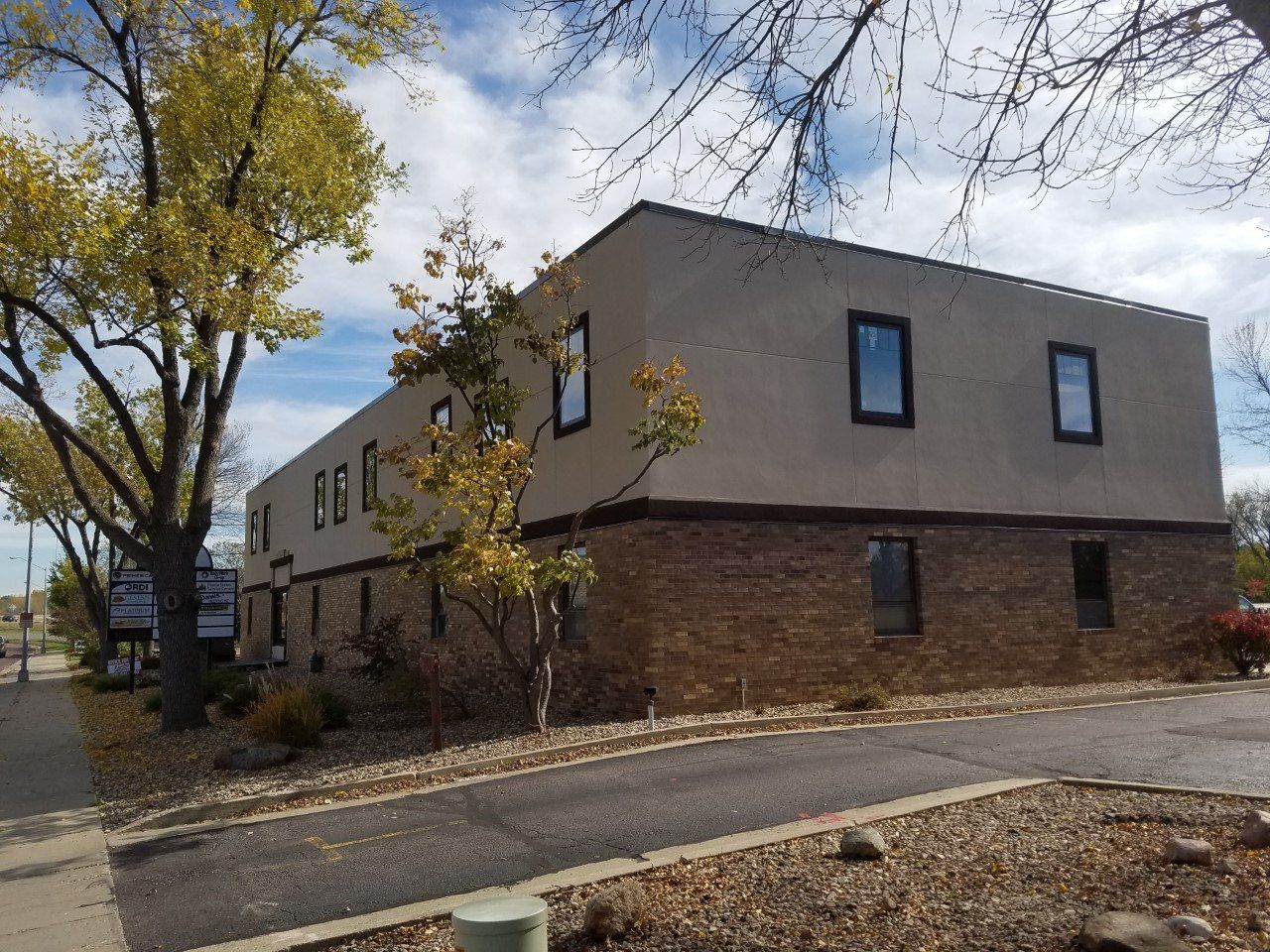 Two-story beige and brown brick commercial building with dark-framed windows, surrounded by trees and a sidewalk.