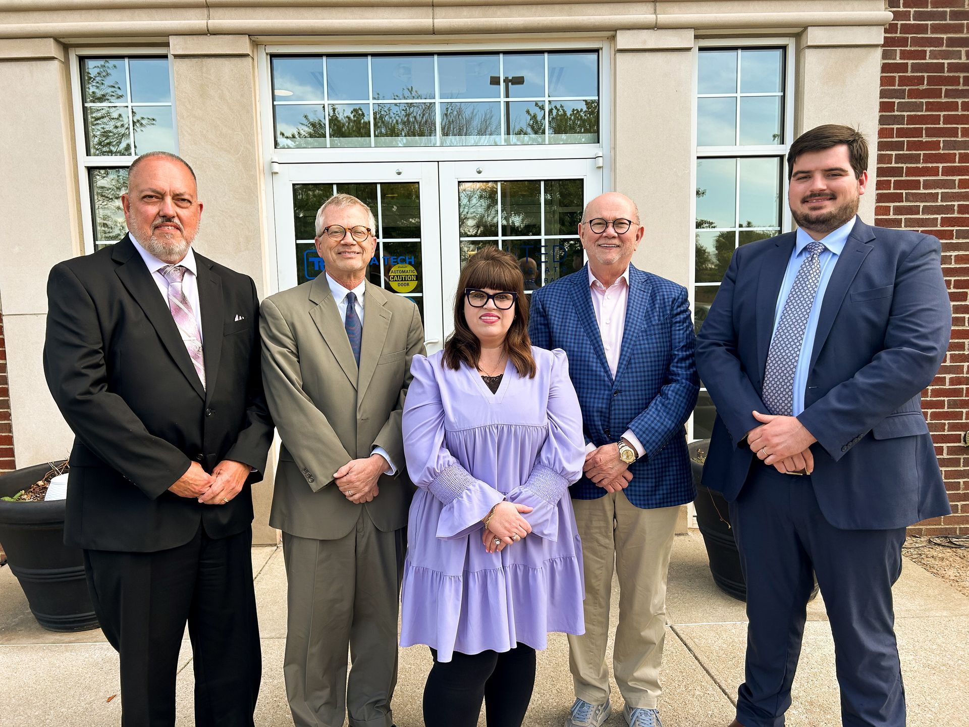 Five people in business attire standing outside a building entrance, posing for a group photo