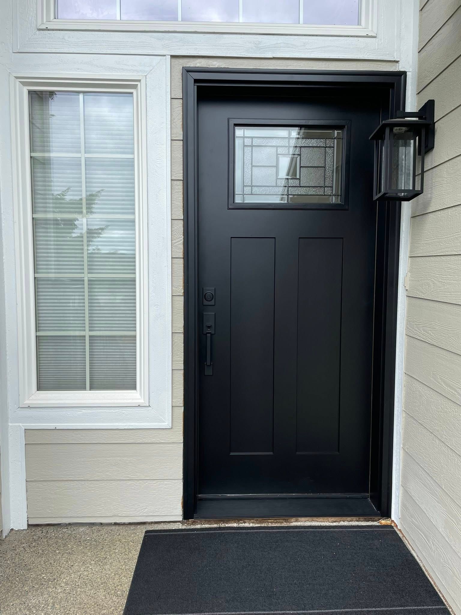 Black front door with glass panel, adjacent white window, and black outdoor light. Beige siding and black doormat are visible.