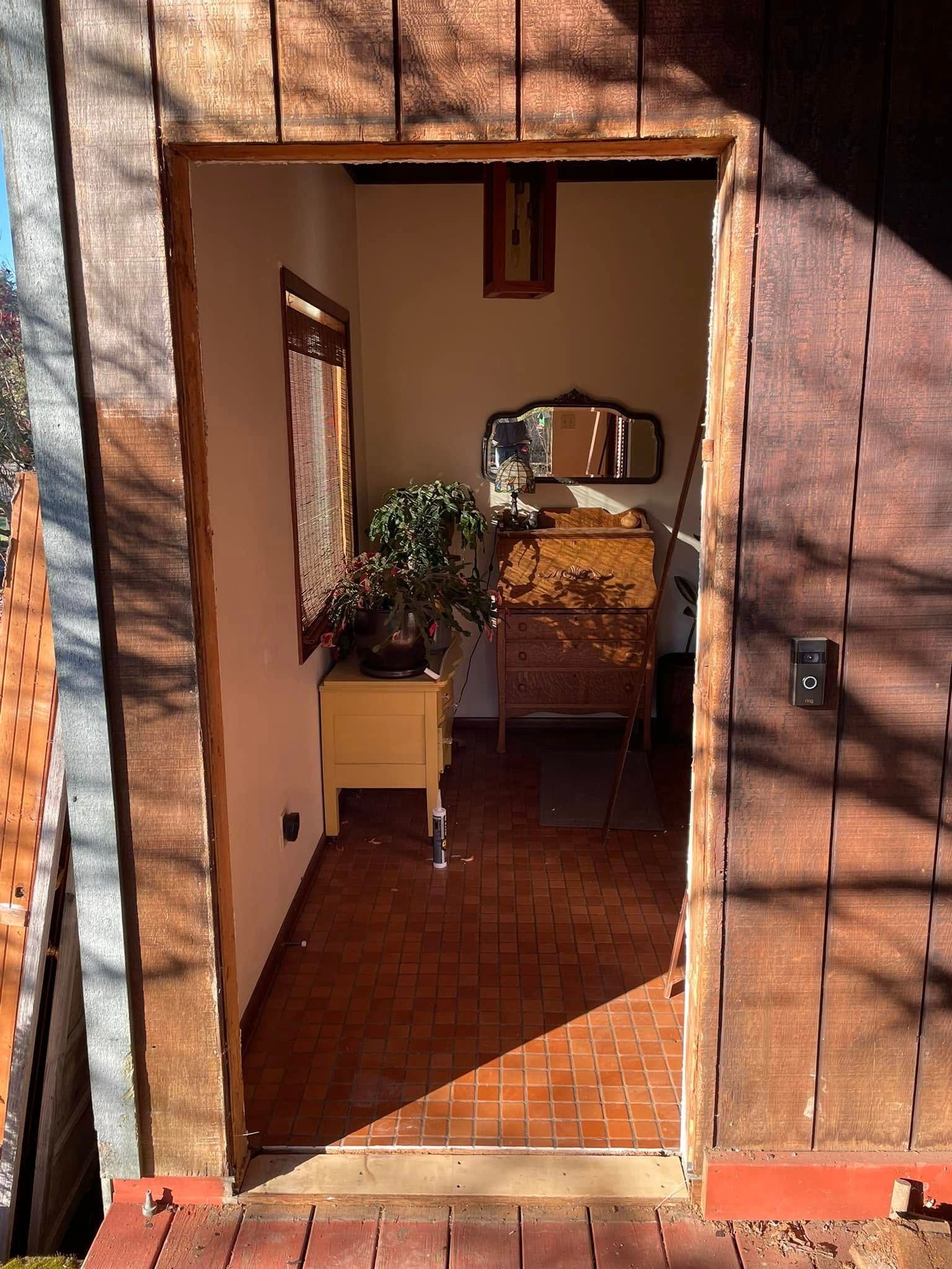 View from doorway into a small room with a dresser, window, and bonsai tree. The room has reddish-brown tiled flooring and wood panel walls.