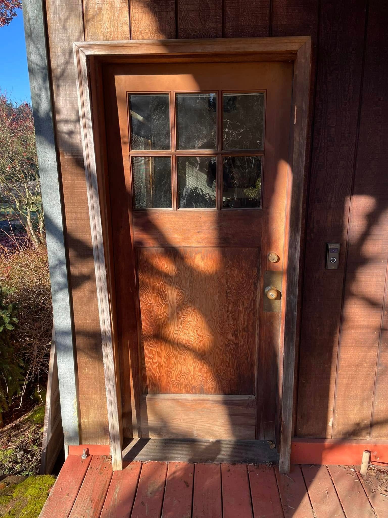 Wooden door with small glass panes, brass handle and doorbell, set into a wooden frame and siding. Shadows cast across it.