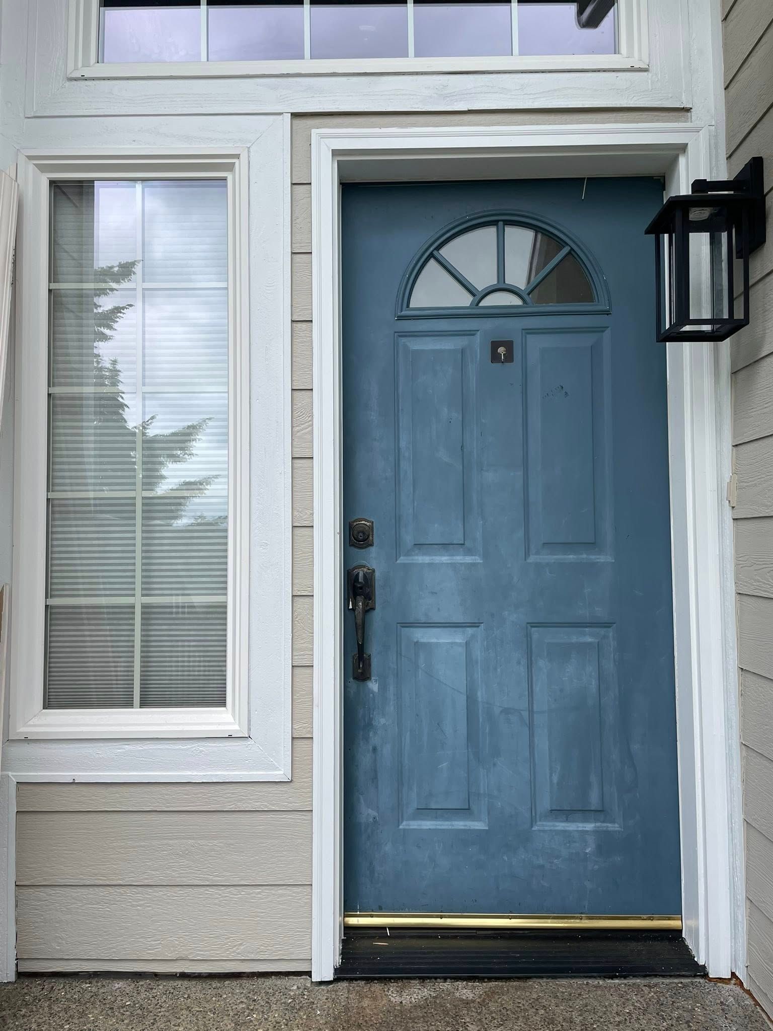 Blue front door with arched glass top, white trim, and black lantern. Beige siding and window with blinds are to the left.