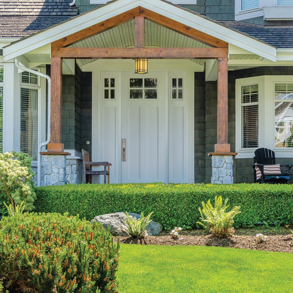 White front door with sidelights and a wooden portico, flanked by stone columns and green shrubbery.