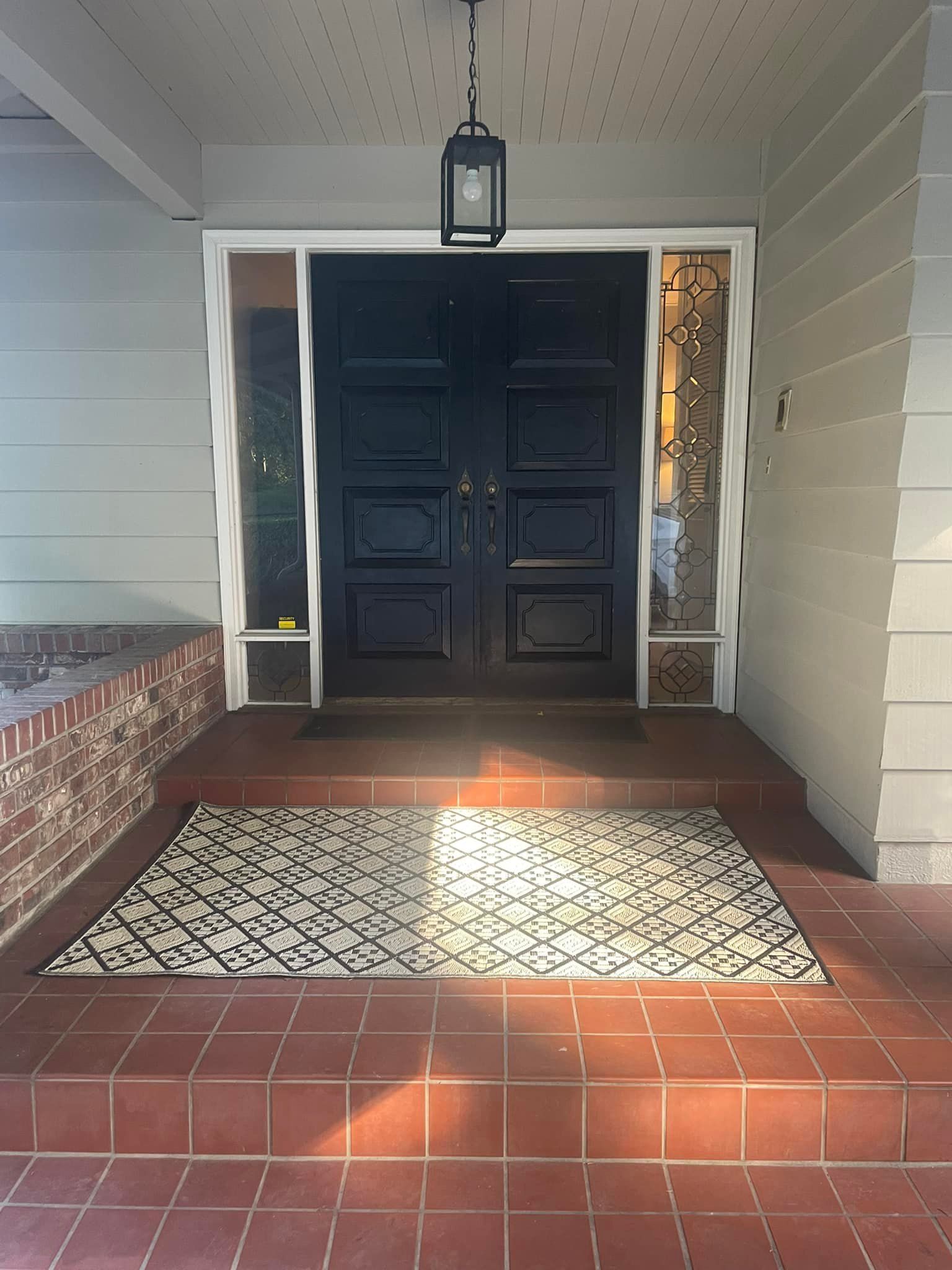 Front entrance with black double doors, sidelights, and brick steps. A patterned rug sits on the top step, with a hanging lantern overhead.