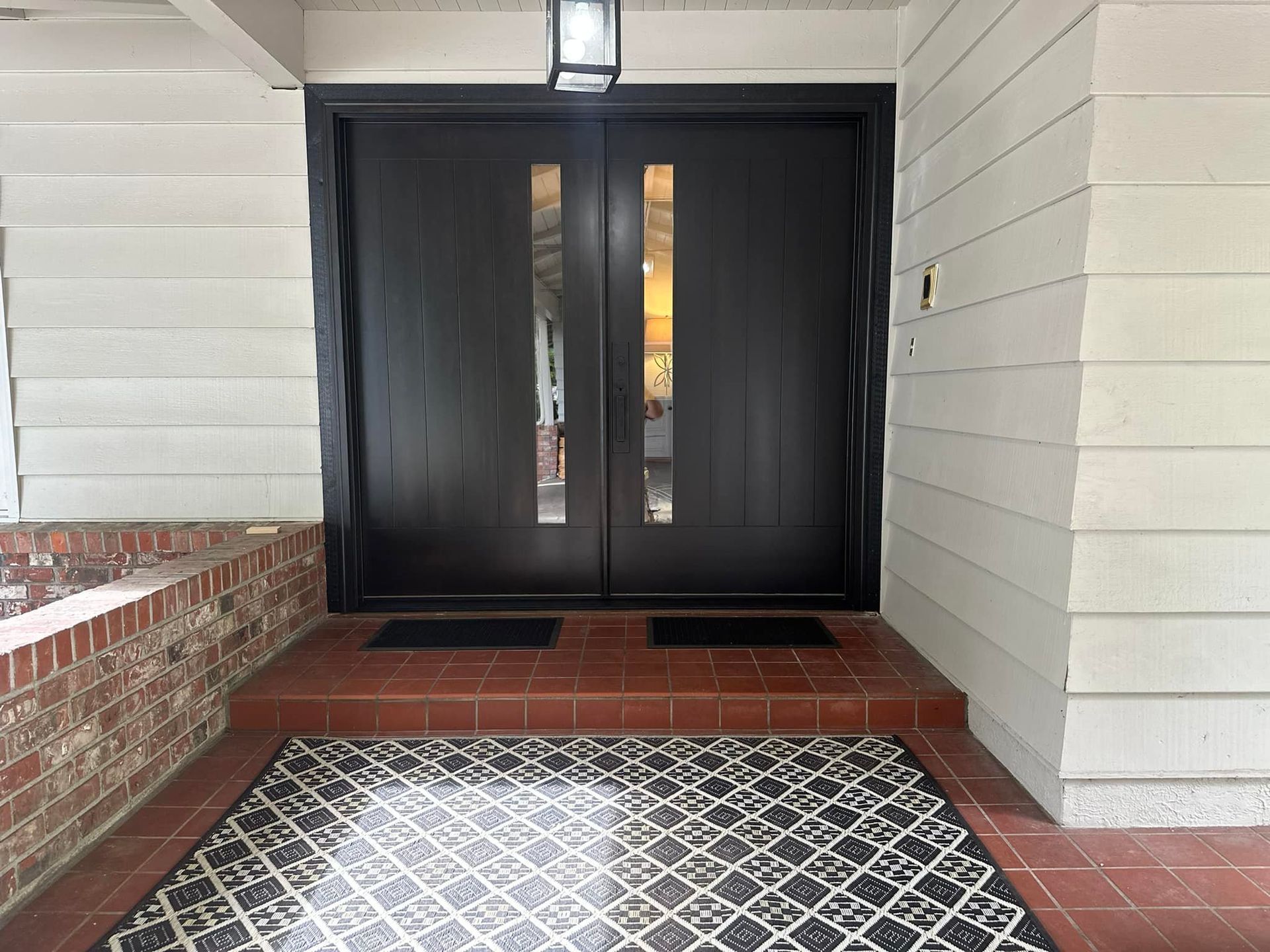 Double black doors with rectangular window panes, set in a white-sided porch, with a patterned rug and brick details.
