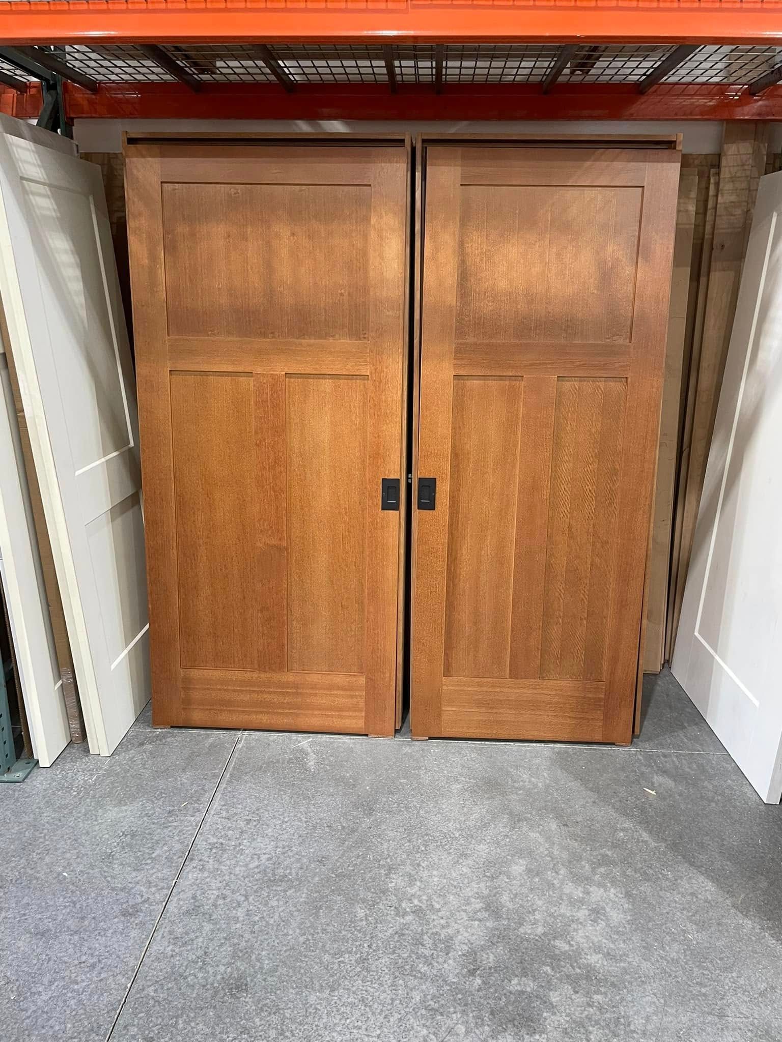 Two wooden sliding doors, medium brown, with rectangular panels and black handles, leaning against a gray floor.