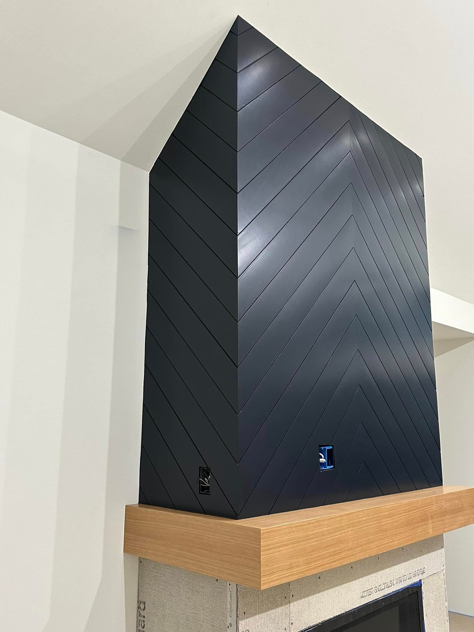 Black angular fireplace surround with chevron pattern over a light wood mantle. The surround is mounted on a light-colored wall.
