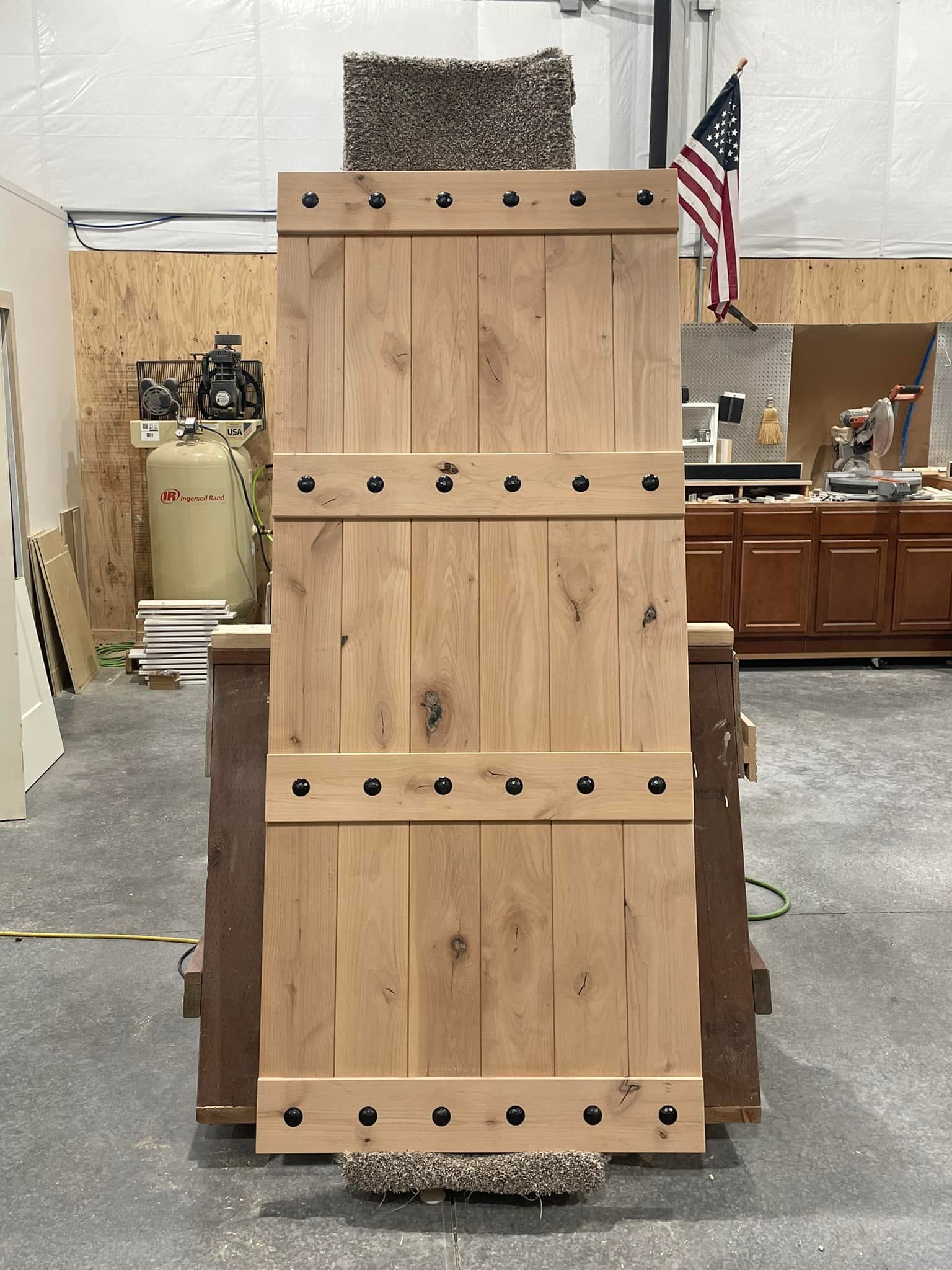 Large, light wooden door with black rivets. It is set on a wooden stand in a workshop, with an American flag in the background.