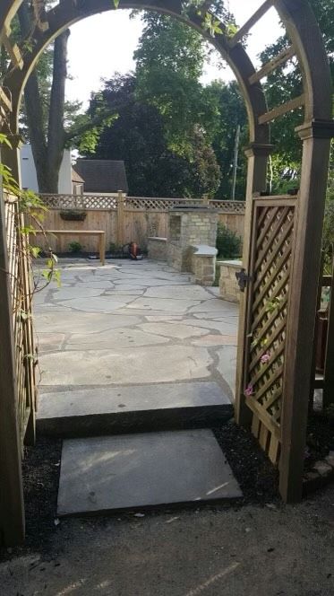 Garden patio seen through a wooden archway with stone paving and a gate on the right.