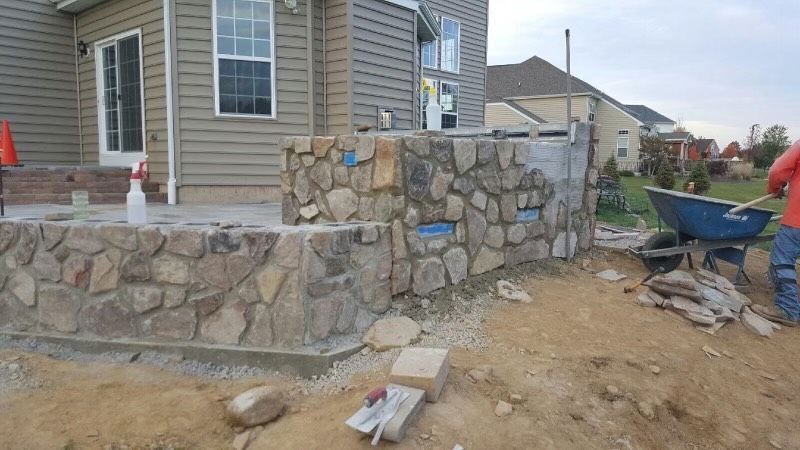 Stone retaining wall under construction beside a beige house, with dirt, tools, and wheelbarrow in the yard.
