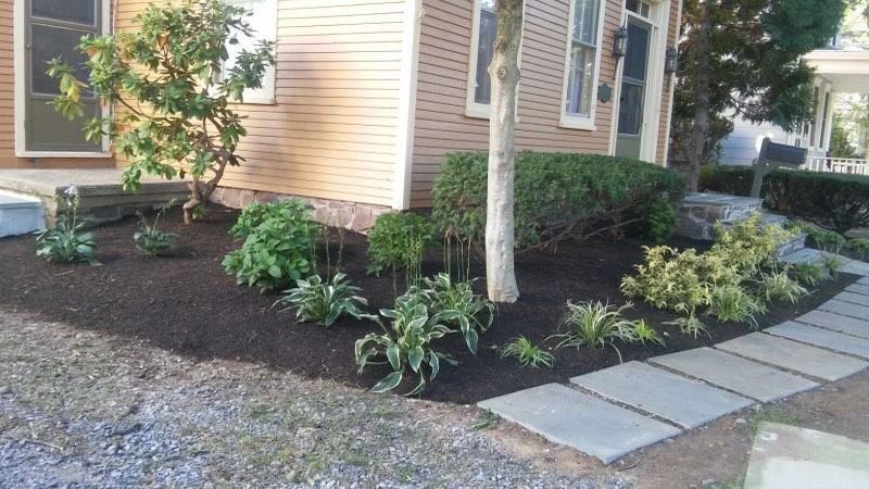Landscaped house corner with mulch beds, shrubs, stepping stones, and a tree beside a gravel driveway