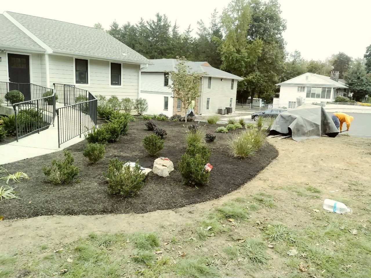 Front yard landscaping with dark mulch, shrubs, rocks, and a house; tarp-covered materials on driveway