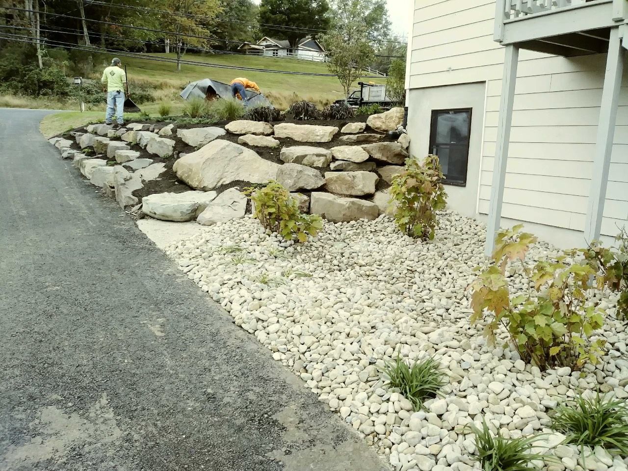 Gravel driveway beside stone retaining wall and white house with landscaped rock garden and shrubs