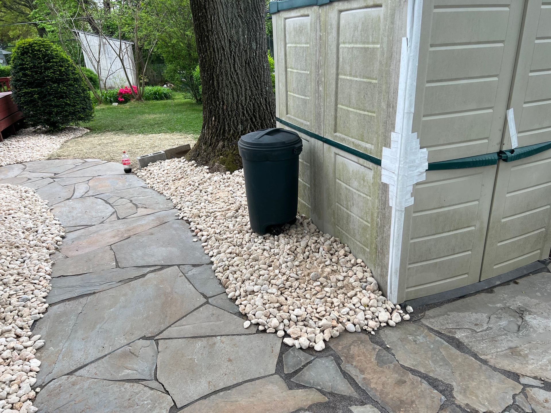 Backyard patio with stone walkway, beige shed, large tree, black trash bin, and white gravel border