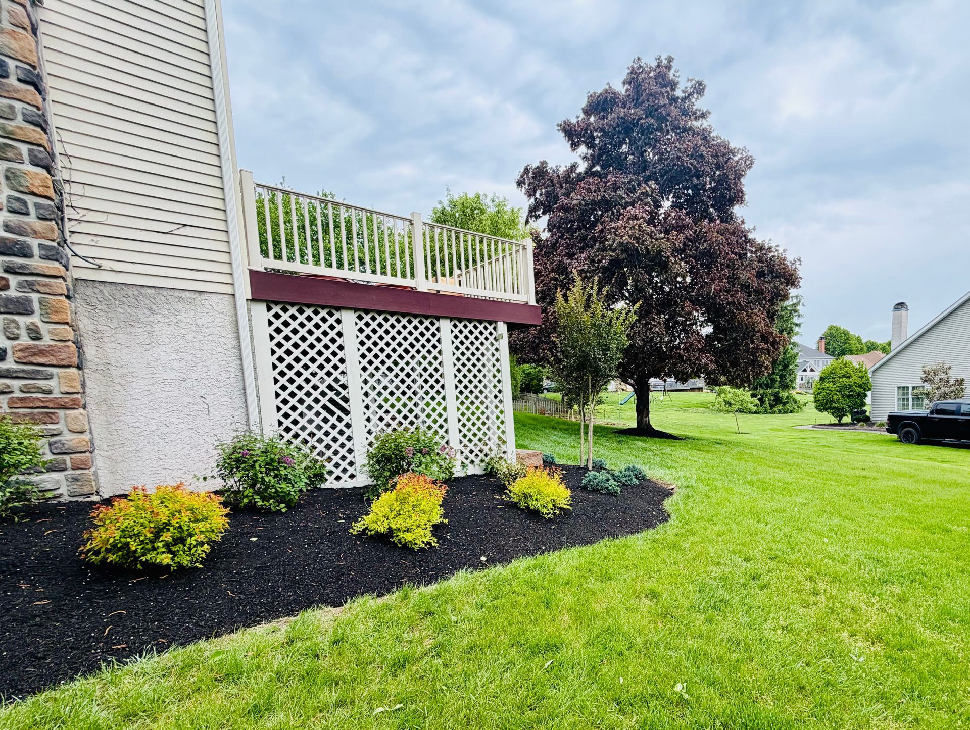 House side yard with landscaped flower bed, dark mulch, and green lawn beside a white deck railing