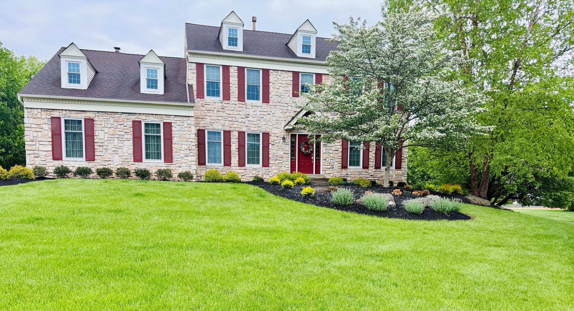 Large brick-and-stone house with red shutters, dormer windows, and a bright green lawn