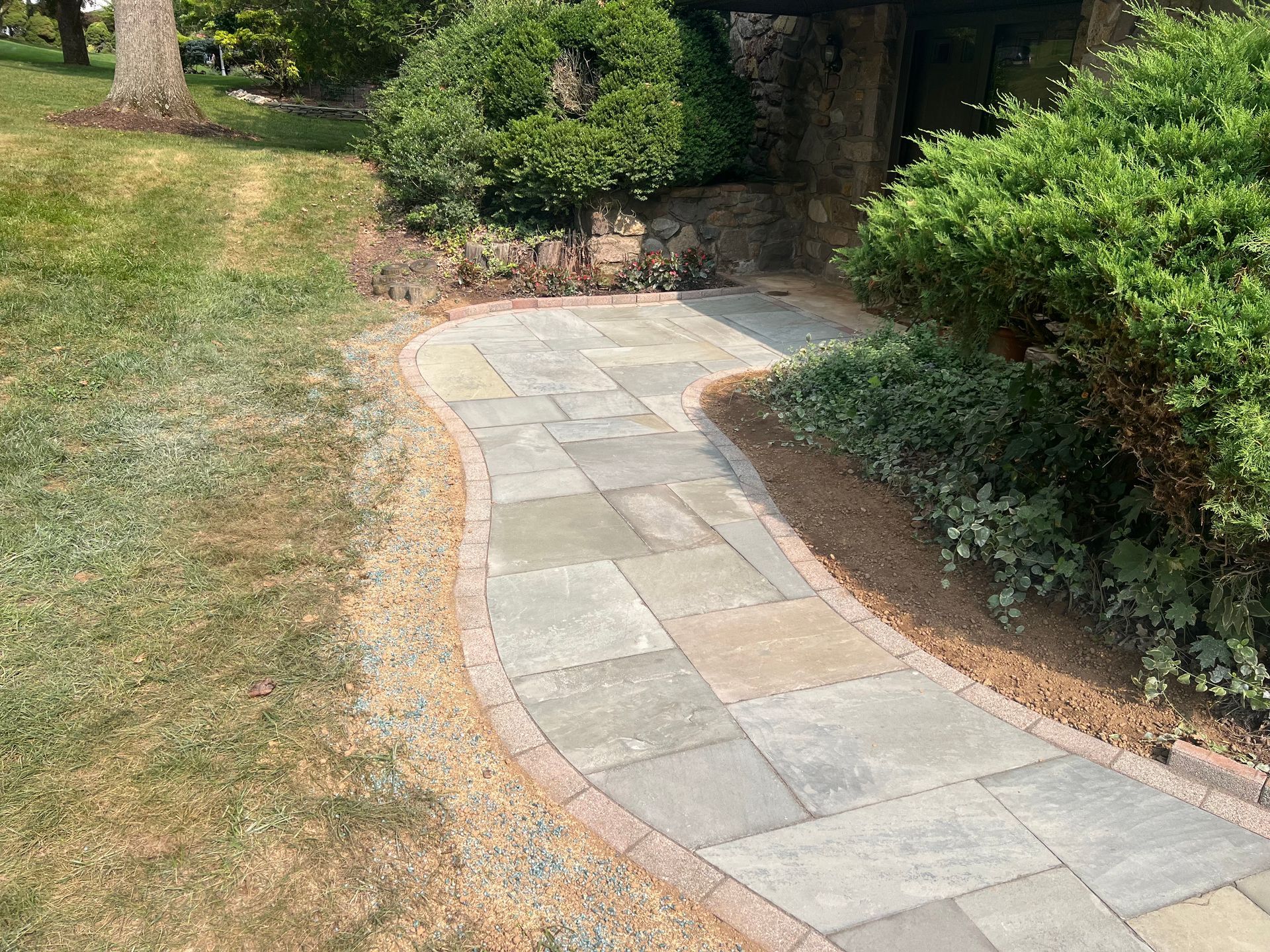 Curving stone garden path bordered by grass and shrubs, leading toward a shaded area.