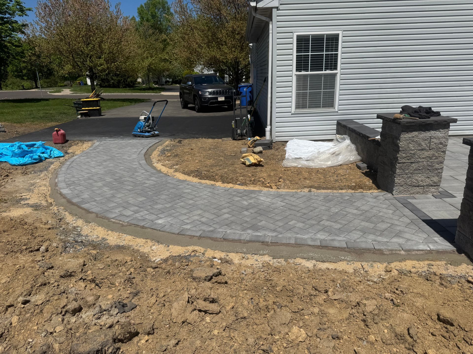 Curved gray paver walkway beside a house, with a car, landscaping, and a driveway in the background.