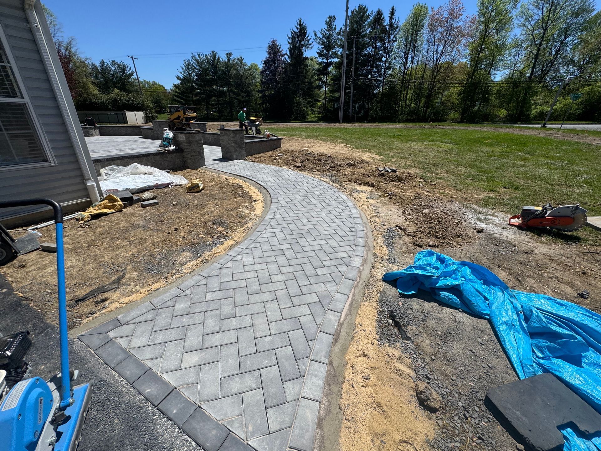Curved gray brick walkway under construction beside a house, with dirt, tools, and blue tarp on a sunny lawn