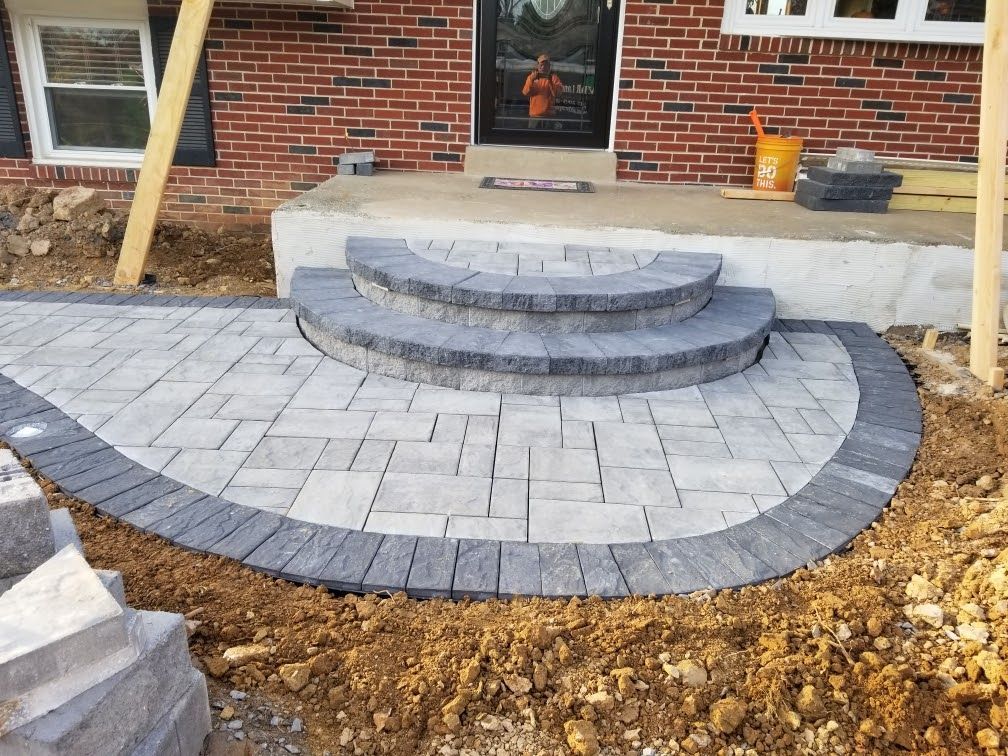 Front steps and semicircular gray paver patio beside a brick house, with unfinished dirt edges